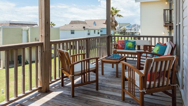 A covered outdoor deck with wooden furniture, including chairs and a table, overlooking a residential area. - Once Upon a Tide by Ryson Vacation Rentals (Galveston)
