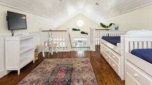 A room with a sloped ceiling, wooden flooring, a television, a crib, a small table, a chair, and a patterned rug. - Once Upon a Tide by Ryson Vacation Rentals (Galveston)