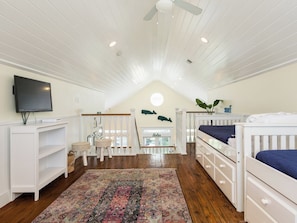 A room with a sloped ceiling, wooden flooring, a television, a crib, a small table, a chair, and a patterned rug. - Once Upon a Tide by Ryson Vacation Rentals (Galveston)