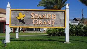 A sign for Spanish Grant with a yellow and black mascot, surrounded by greenery and palm trees. - Once Upon a Tide by Ryson Vacation Rentals (Galveston)