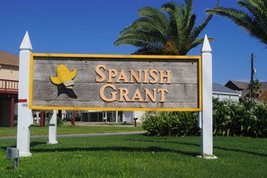 A sign for Spanish Grant with a yellow and black mascot, surrounded by greenery and palm trees. - Once Upon a Tide by Ryson Vacation Rentals (Galveston)