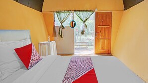A bedroom with a bed, pillows, a bedside table, a small table, and a door leading to an outdoor area. - OYO 90474 Caravan Camp (Batu)