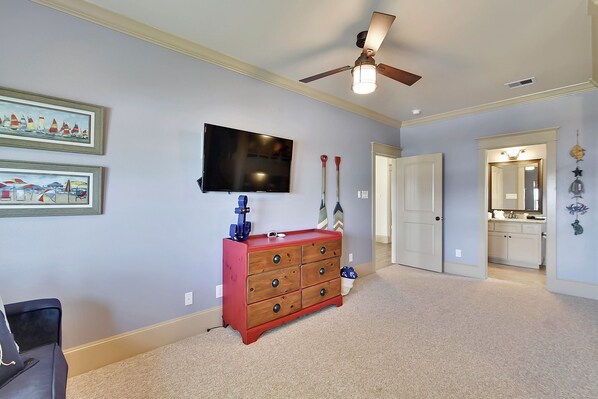 A room with a ceiling fan, a TV mounted on the wall, a wooden dresser, and a door leading to a bathroom.