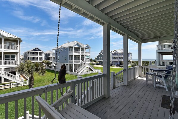 A covered porch with a swing chair, a view of beachfront houses, and a palm tree.