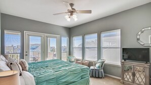 A bedroom with a bed, a ceiling fan, a TV, and large windows. - Mutiny on the Bounty by Ryson Vacation Rentals (Galveston)