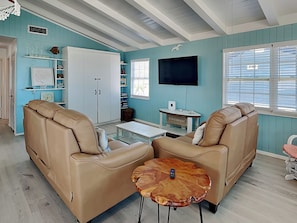 A living room with two beige leather sofas, a wooden coffee table, a flat-screen TV, and a bookshelf with decorative items. - Paradise Found by Ryson Vacation Rentals (Galveston)