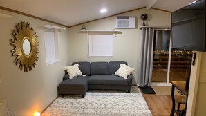 Un salon avec un canapé d’angle gris, un tapis à motifs, un miroir avec un cadre doré et un ventilateur de plafond.