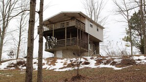 Une cabane dotée d’un balcon, située sur une colline, entourée d’arbres et de zones enneigées.