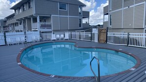A swimming pool with a wooden deck, surrounded by white fences and chairs, and adjacent to a building with a balcony.