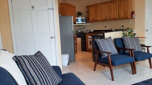 Una sala de estar con un sofá y dos sillones, una cocina con armarios de madera y un microondas, y una puerta blanca.