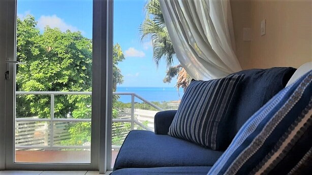 Habitación con vista al océano y vegetación, puerta de vidrio corredera y sofá azul con almohadones a rayas.