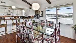 Un área de comedor con una mesa de vidrio rodeada de sillas estampadas, armarios blancos y un ventanal con vista a un cuerpo de agua.