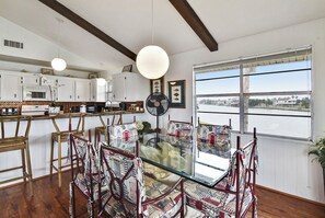 A dining area with a glass table surrounded by patterned chairs, white cabinets, and a large window with a view of a body of water.