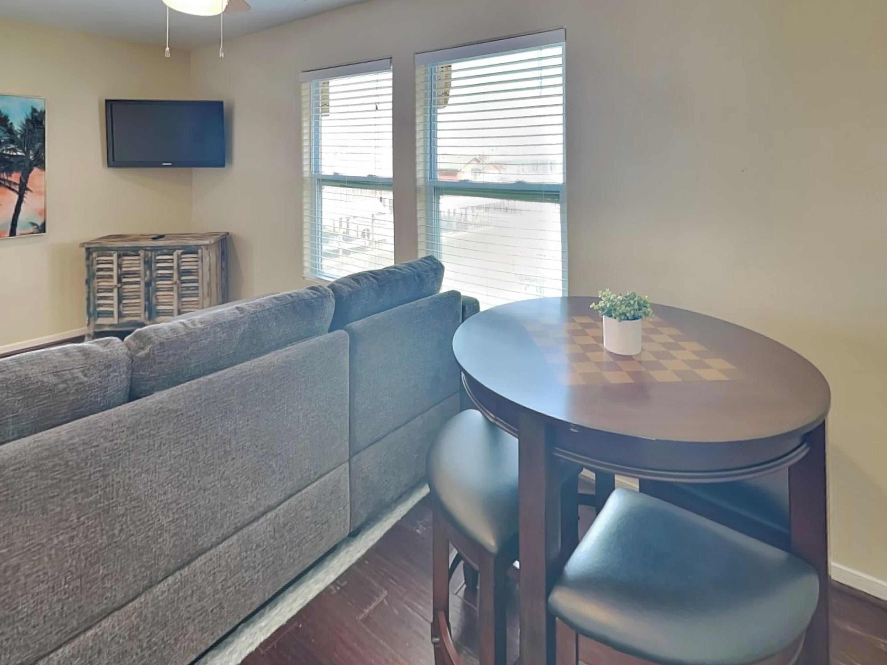 A living room with a gray sofa, a round wooden table with a chessboard, and a TV mounted on the wall.