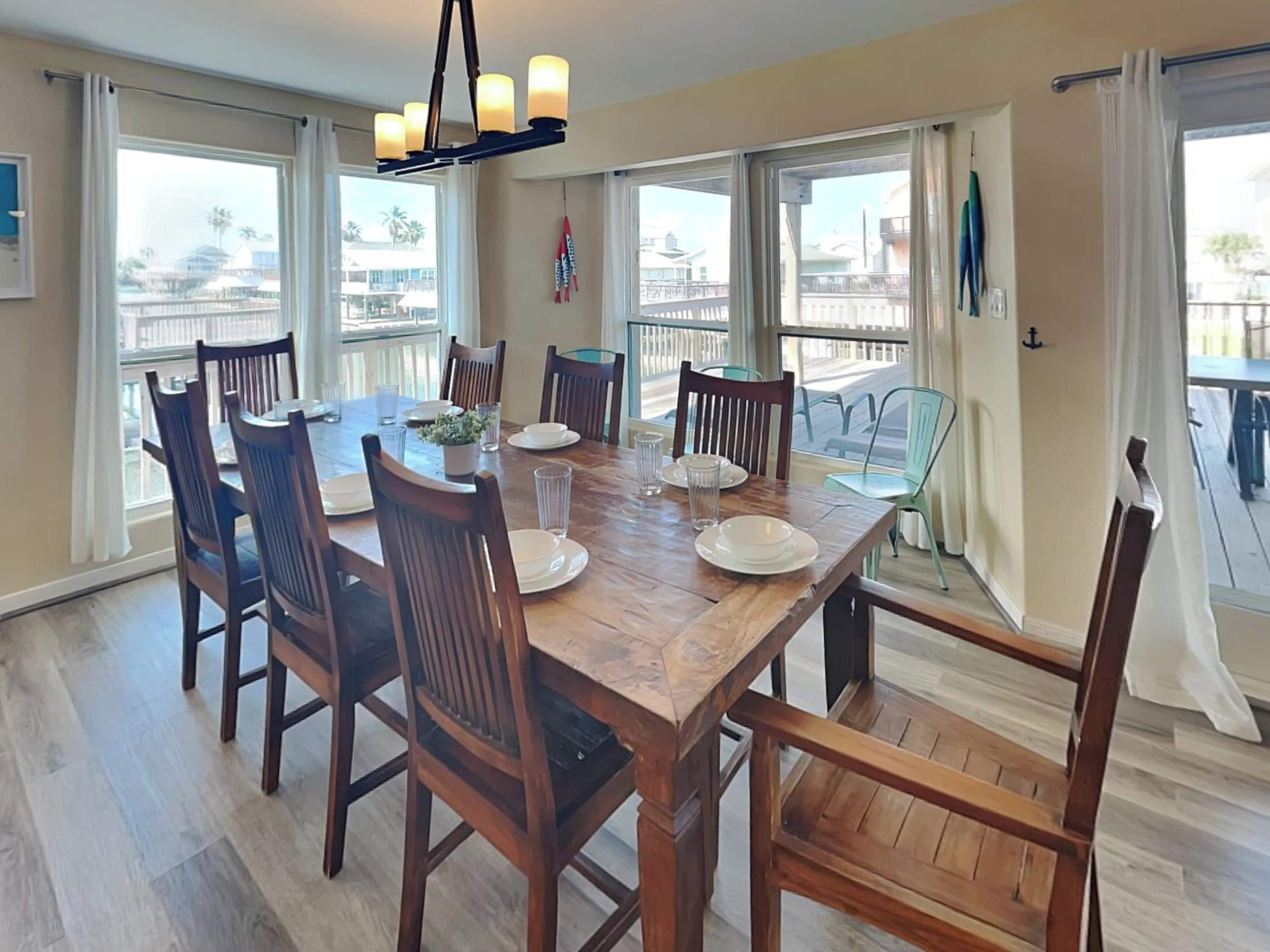 A dining room with a wooden table set for six, surrounded by chairs, large windows, and a view of the outside.