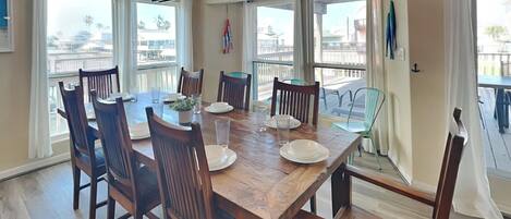A dining room with a wooden table set for six, surrounded by chairs, large windows, and a view of the outside.
