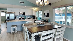 A modern kitchen with white cabinetry, a central island, and a dining area with white chairs. - Reel Paradise by Ryson Vacation Rentals (Galveston)