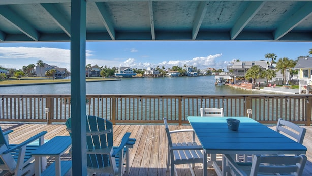 A wooden deck with blue chairs and a table overlooking a body of water with houses in the background. - Reel Paradise by Ryson Vacation Rentals (Galveston)