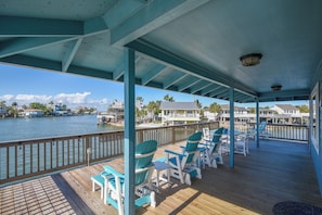 A covered deck with blue Adirondack chairs and a view of the water and buildings. - Reel Paradise by Ryson Vacation Rentals (Galveston)