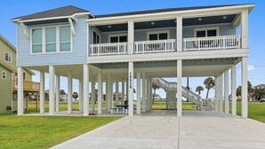 Una casa frente a la playa de dos pisos, con un porche cubierto, un balcón y una escalera que conduce al nivel superior.