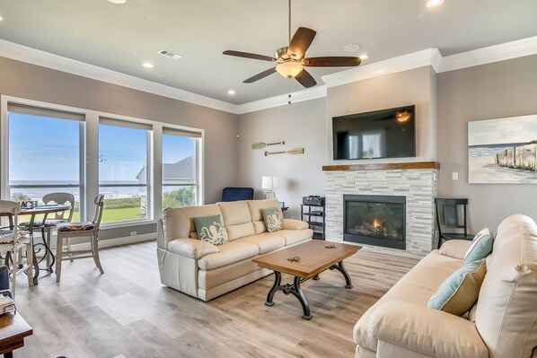A spacious living room with a fireplace, large windows, and a ceiling fan. - Salty Blue Mermaid by Ryson Vacation Rentals (Galveston)