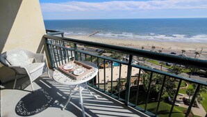 Varanda com vista para a praia, uma cadeira de vime, uma mesa pequena com um tabuleiro e um corrimão.