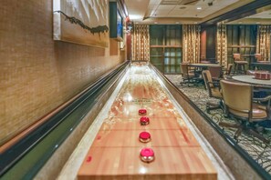 A long, narrow pool table with a polished surface and red markers, surrounded by chairs and tables in a room with patterned carpet and decorative walls.