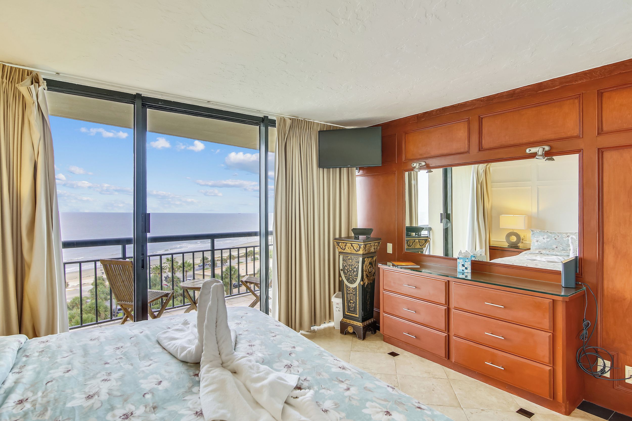 A bedroom with a bed, a dresser, a TV, and a balcony with a view of the sea.