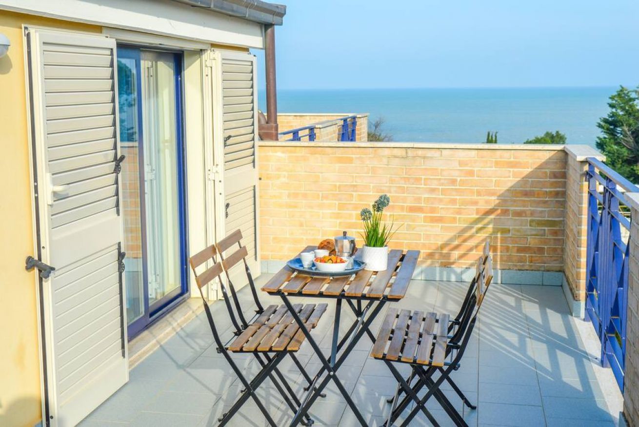 A balcony with a table set for two, a potted plant, and a view of the sea.