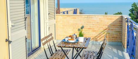 A balcony with a table set for two, a potted plant, and a view of the sea.
