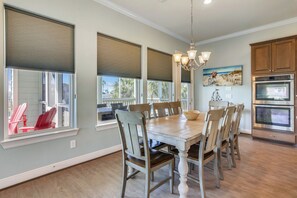 A dining area with a wooden table and chairs, large windows with blinds, a chandelier, and a kitchen with stainless steel appliances.