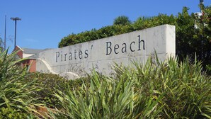 A sign reading "Pirates' Beach" with surrounding greenery and a building in the background. - Sandpiper Treasure by Ryson Vacation Rentals (Galveston)