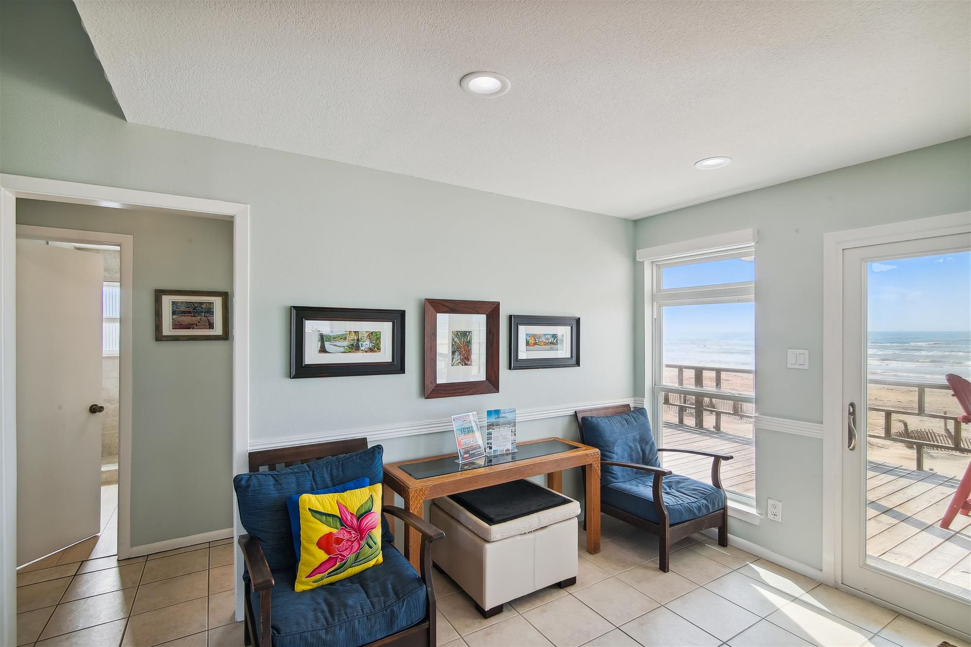 A living room with a view of the beach, a dining area with a table and chairs, and a door leading to another room.