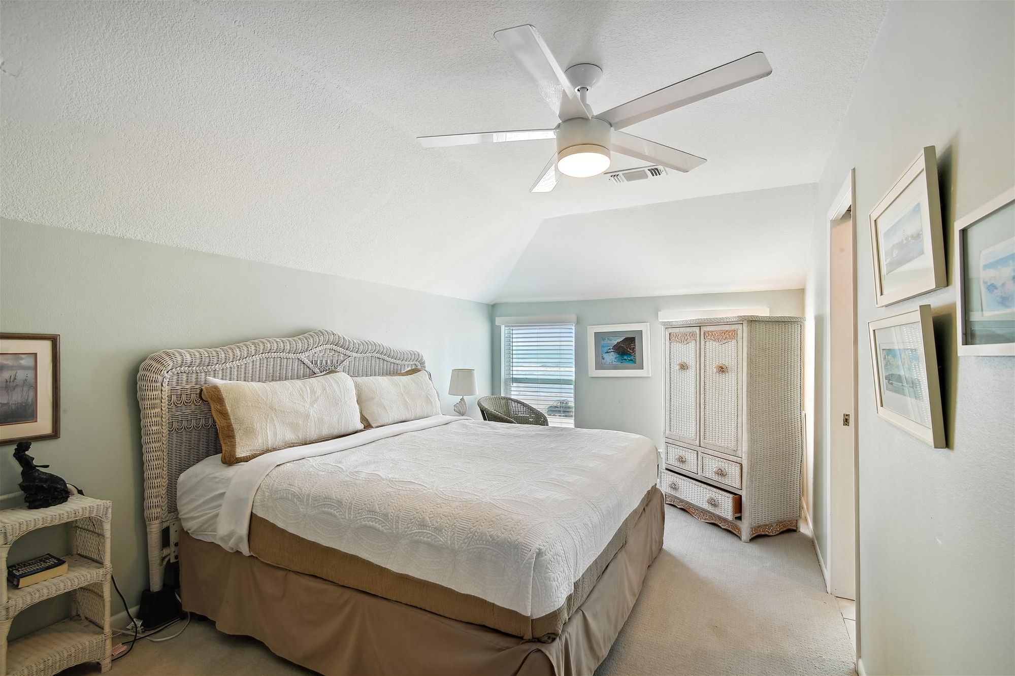 A bedroom with a bed, a ceiling fan, a wicker chair, a bedside table, and a wardrobe.