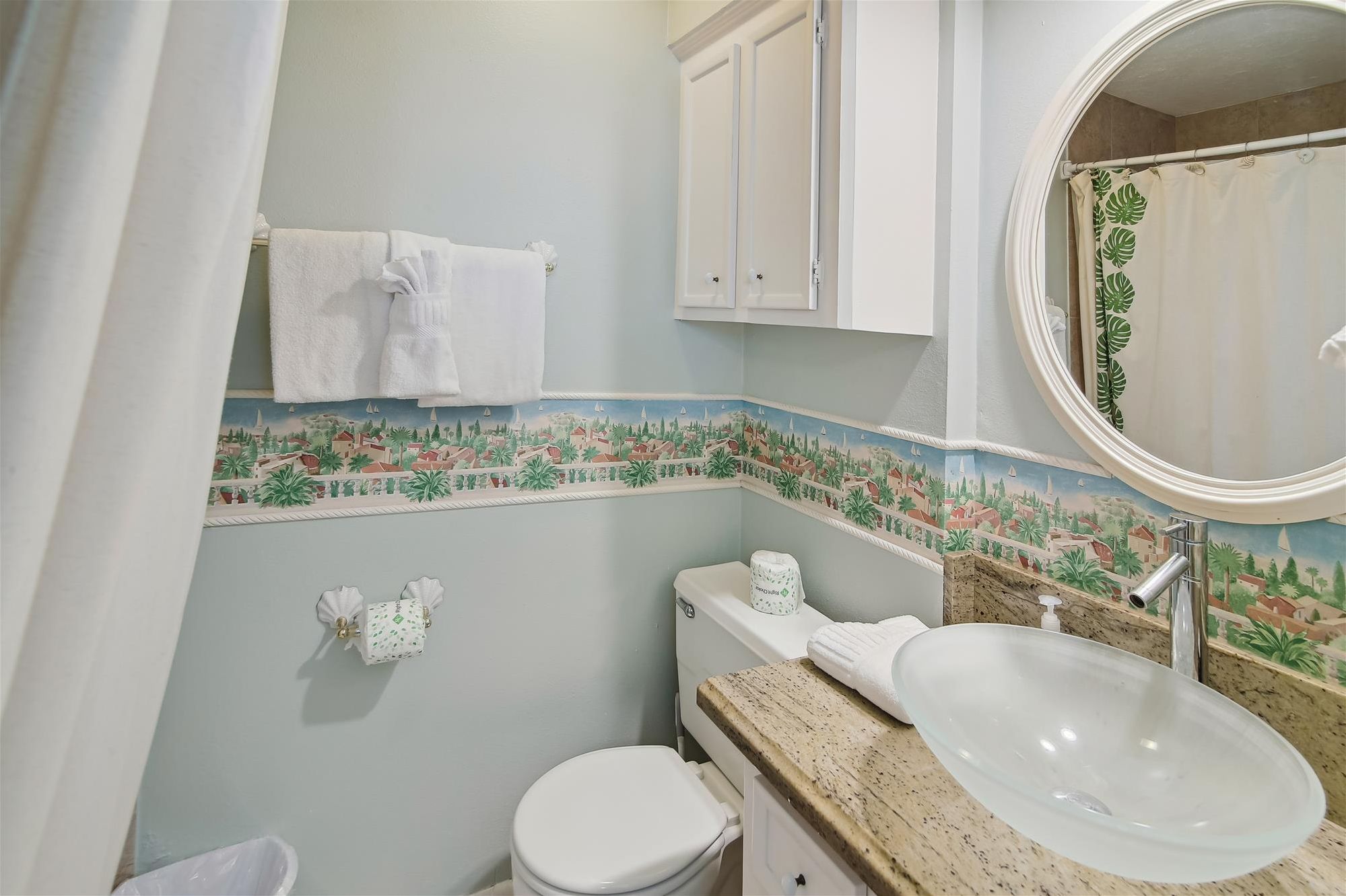 Une salle de bain avec des toilettes blanches, un lavabo avec un miroir et un rideau de douche orné d’un motif de feuilles.