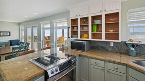 A kitchen with a built-in stove, microwave, and granite countertop. There are open shelves with dishes and a view of the ocean through large windows. - Sea Loft by Ryson Vacation Rentals (Galveston)