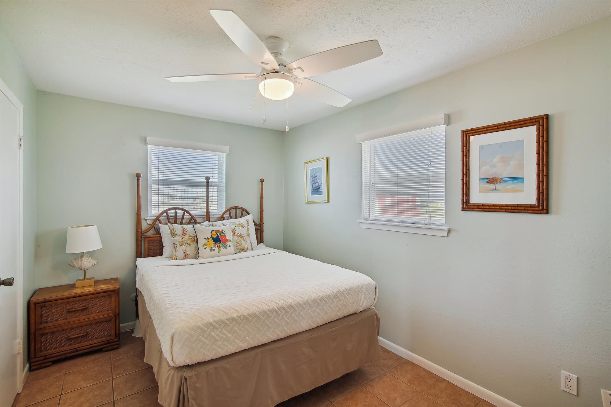 A bedroom with a bed, a nightstand, a lamp, a ceiling fan, and two windows with blinds.