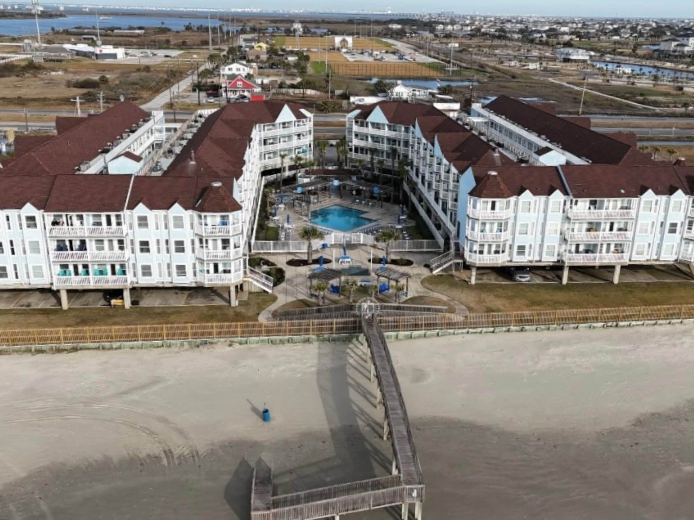 Un hotel frente a la playa con piscina central, varios balcones y un paseo marítimo que da a la playa.