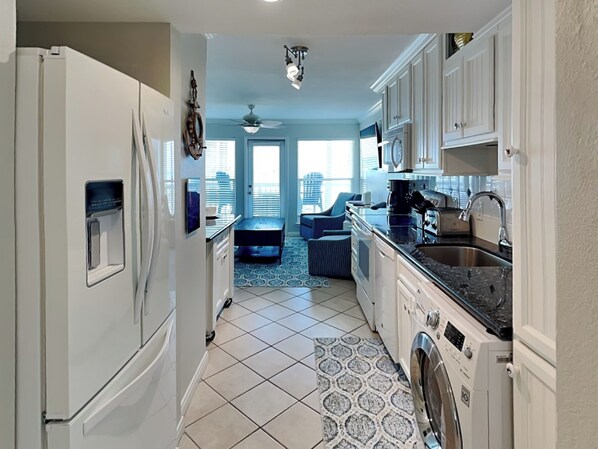 A compact kitchen with a refrigerator, washing machine, and a dining area with a blue chair. - Beachcomber by Ryson Vacation Rentals (Galveston)