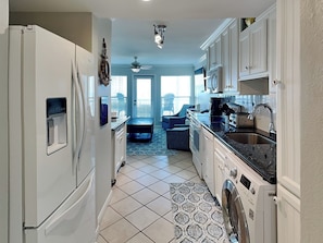 A compact kitchen with a refrigerator, washing machine, and a dining area with a blue chair. - Beachcomber by Ryson Vacation Rentals (Galveston)