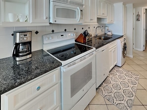 A modern kitchen with white cabinetry, a built-in microwave, a stove, a coffee maker, a sink, and a washing machine. - Beachcomber by Ryson Vacation Rentals (Galveston)
