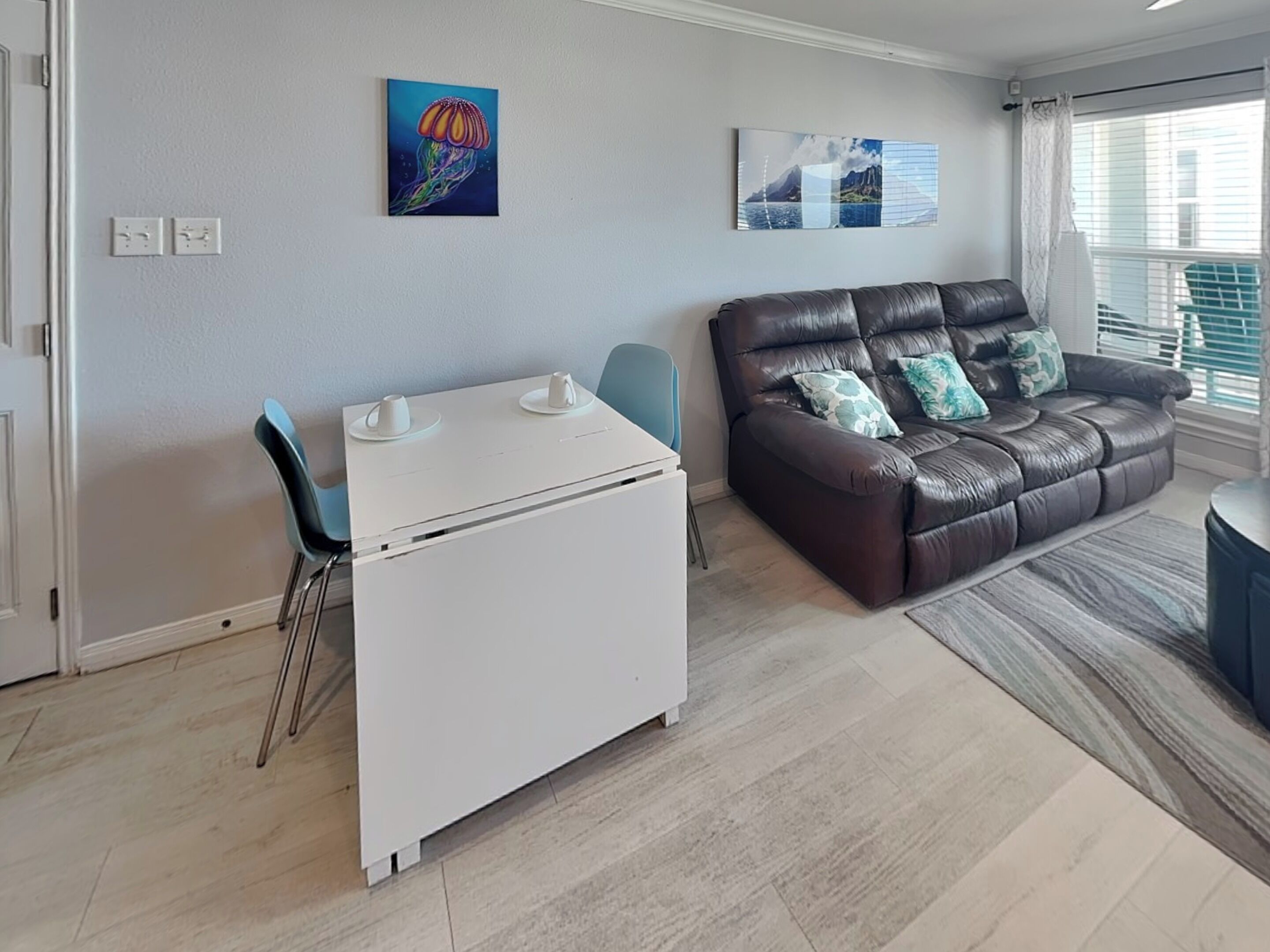 A living room with a brown leather sofa, a white coffee table, and a blue chair. There are two paintings on the wall. - A Sunny Seahorse by Ryson Vacation Rentals (Galveston)