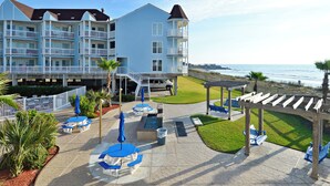 Un hotel frente a la playa con varios balcones, mobiliario exterior y zona de piscina.