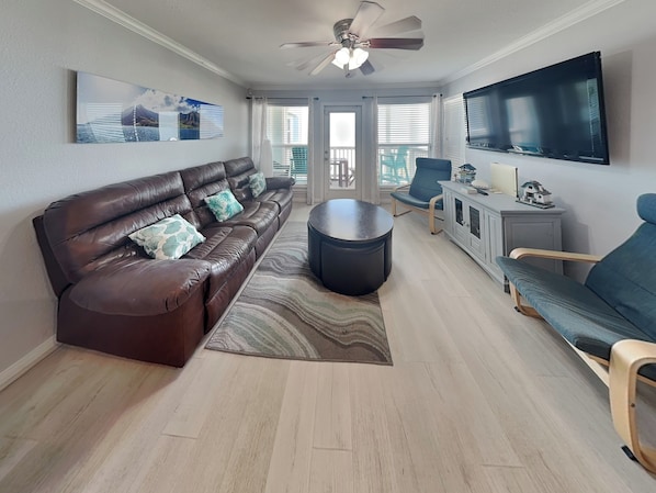 A living room with a brown leather sofa, a round coffee table, a blue armchair, a TV, and a ceiling fan. - A Sunny Seahorse by Ryson Vacation Rentals (Galveston)
