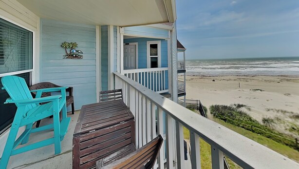 A balcony with a view of the beach, a wooden table, turquoise chairs, and a small plant decoration.