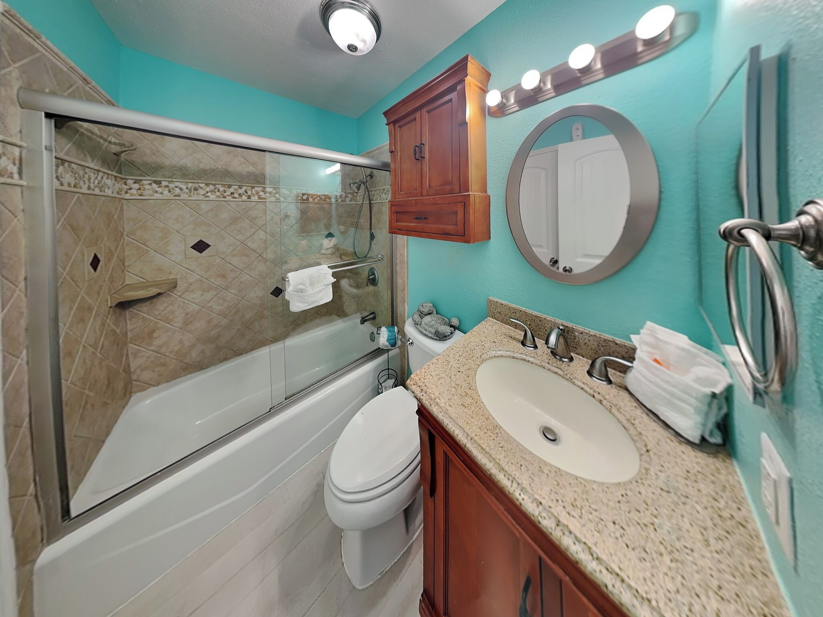 A bathroom with a glass-enclosed shower, a toilet, a sink with a granite countertop, wooden cabinets, and a mirror with lights.