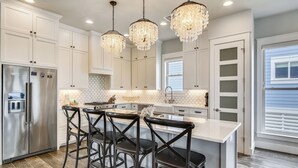 A modern kitchen with white cabinetry, a stainless steel refrigerator, a central island with bar stools, and decorative crystal chandeliers. - Skin and Bones by Ryson Vacation Rentals (Galveston)