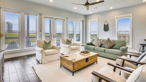 A spacious living room with a large window, a ceiling fan, and a wooden coffee table. - Skin and Bones by Ryson Vacation Rentals (Galveston)