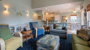 Un salón espacioso con un amplio sofá azul, un sillón verde y un otoman decorado. Se observa una zona de comedor con una mesa y sillas, y una cocina al fondo.