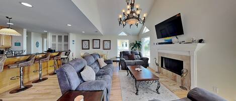 A spacious living room with a large sofa, a coffee table, and a fireplace. There is a television mounted above the fireplace and a chandelier hanging from the ceiling.